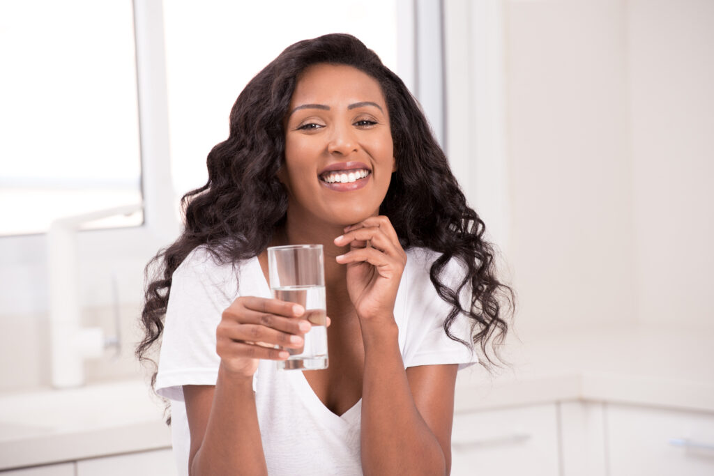 Woman wearing white v-shape shirt, drinking mineral water, sitting at kitchen bar and looking over the camera with a friendly smile.