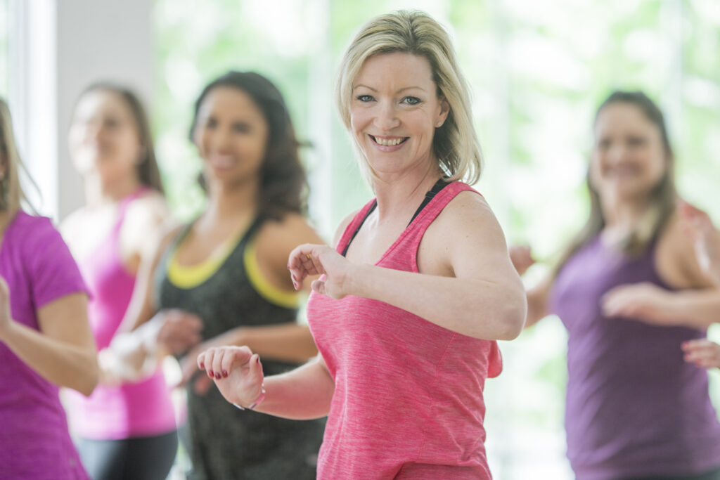 A multi-ethnic group of adult women are taking a dance fitness class indoors.