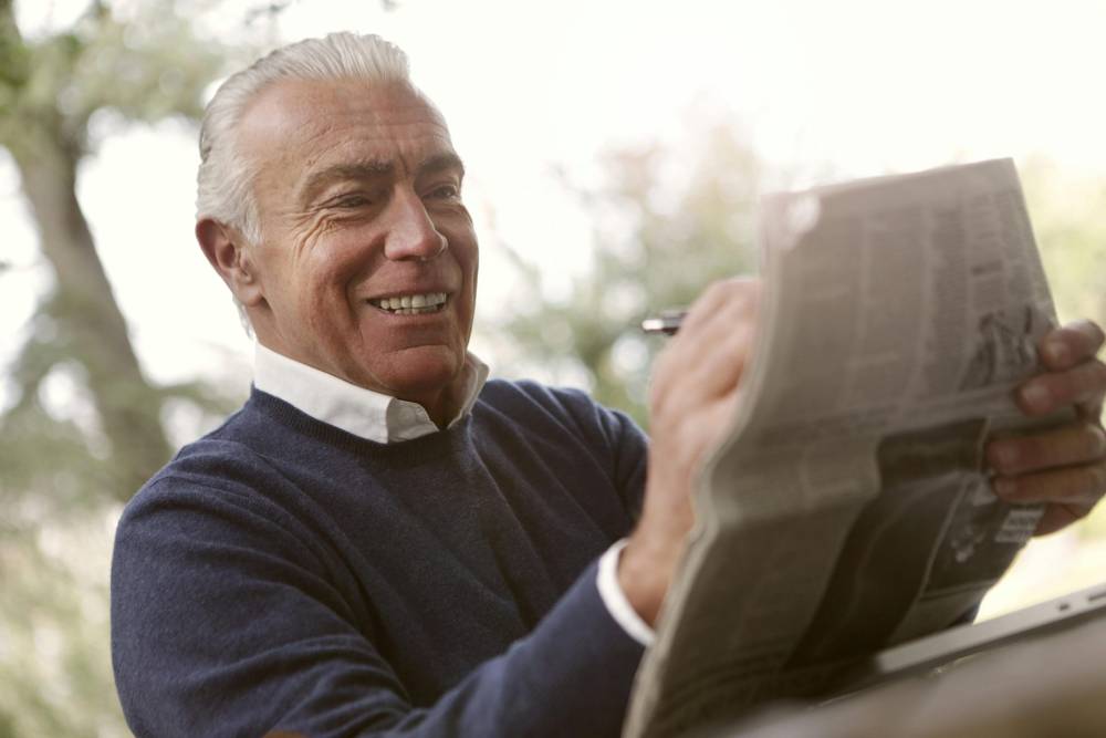 Elderly man smiling and reading the newspaper outdoors, enjoying his leisure time.